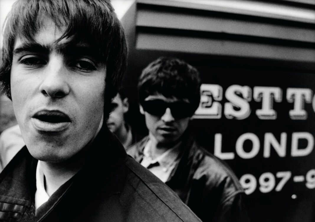 Im Vordergrund blickt Liam Gallagher arrogant in die Kamera, im Hintergrund schaut Noel Gallagher mit Sonnenbrille über Liams Schulter.