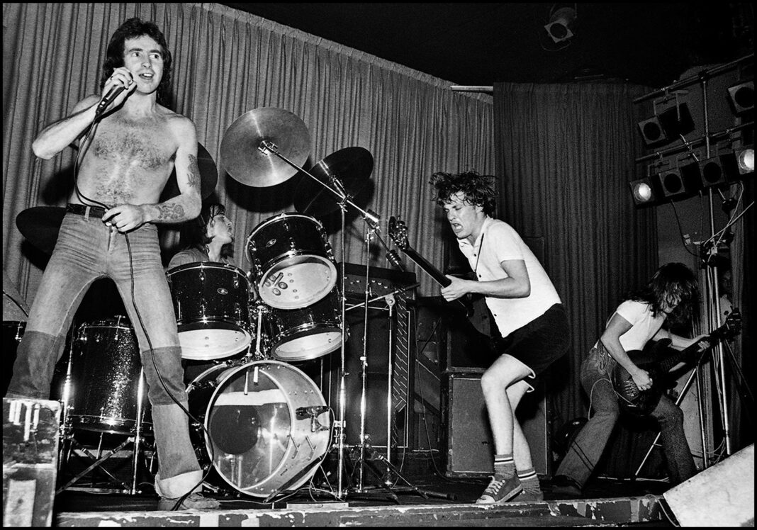 Bon Scott steht mit freiem Oberkörper und Mikrofon in der Hand auf der Bühne, neben ihm spielen Angus und Malcolm Young mit wehenden Haaren Gitarre.