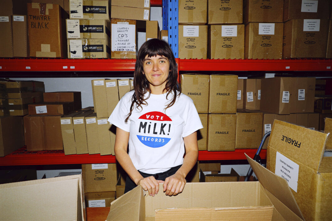 Courtney Barnett steht in einem Lagferraum vor einem KArton und trägt ein T-Shirt mit der Aufschrift 