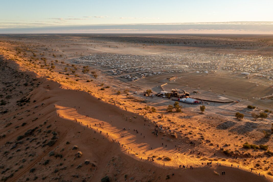 Aus der Ferne sieht man vorne Menschen auf einer rot glühenden Outback-Wüstensand-Düne, im Hintergrund stehen Fahrzeuge im Halbkreis um eine große Bühne.